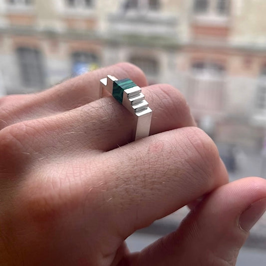 Bague carrée en argent brossé, au centre un cube de malachite est enchâssé entre deux éléments inversés en forme d'escalier. Bijou sur une main d'homme.