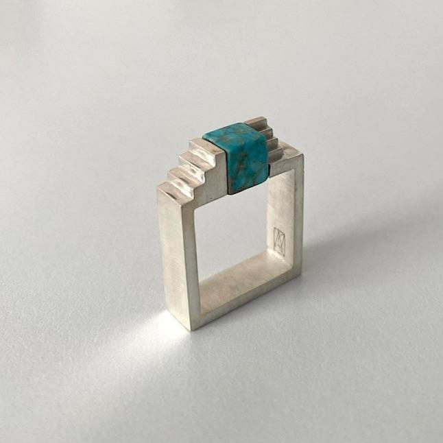 Bague carrée en argent brossé, au centre turquoise  taillée en cube est enchâssée entre deux éléments inversés en forme d'escalier.