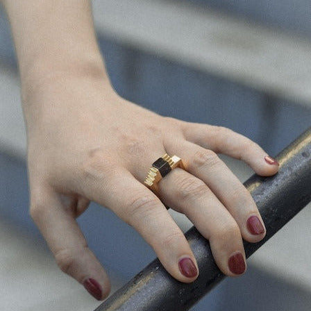 Bague carrée en vermeil, au centre un onyx noir taillée en cube est enchâssé entre deux éléments inversés en forme d'escalier. Bijou présenté sur une main de femme.