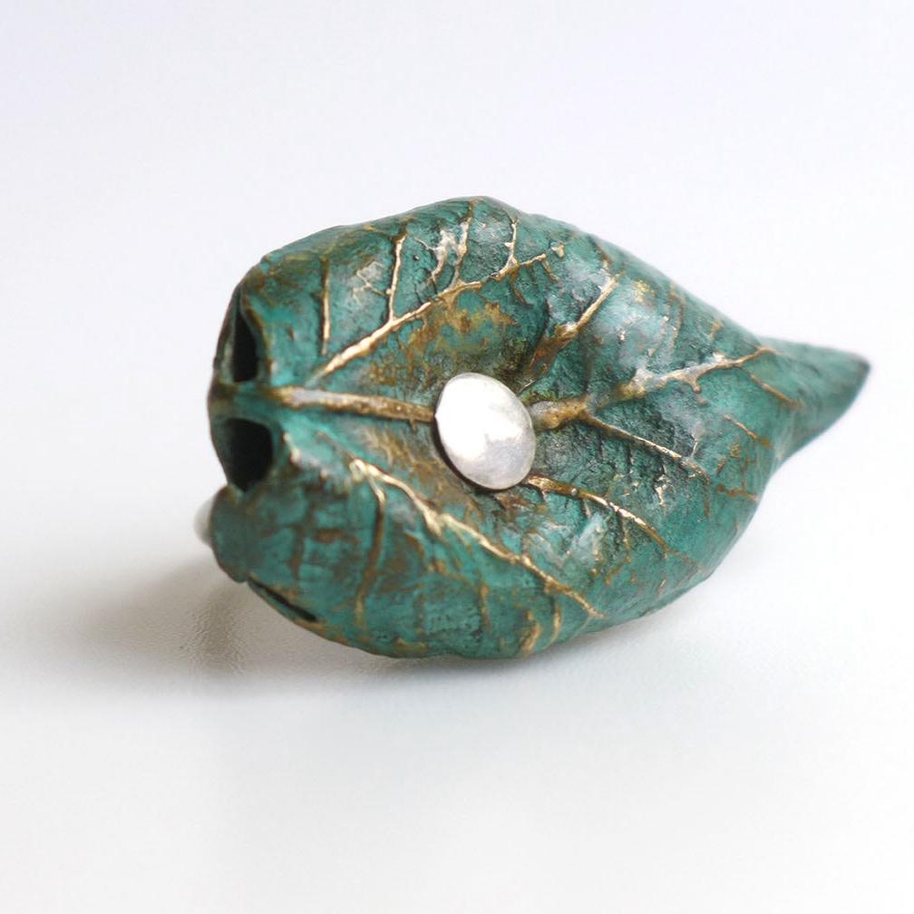 Cette bague est née du moulage de véritables feuilles d'arbres. Réalisée en bronze patiné au vert-de-gris, elle révèle des détails organiques. L'ensemble est présenté sur un anneau en argent forgé. 