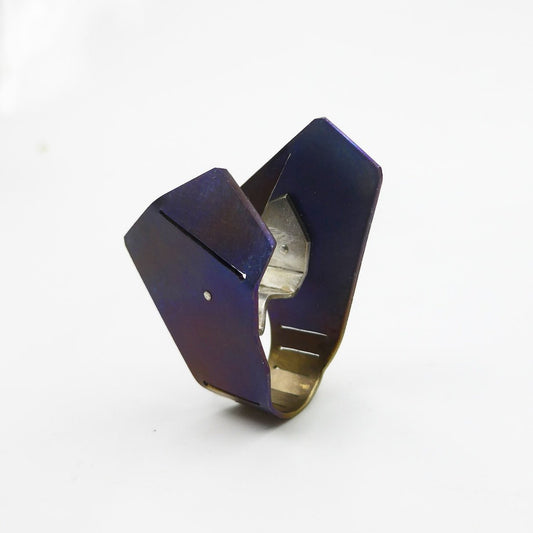 Bague construite comme une sculpture, à partir d'une plaque de titane pliée, finement sciée, puis rivetée à un plaque d'argent brossé. La surface du métal légèrement brossée est de couleur bleu foncé, presque violet. Ce bijou aérienne ressemble à deux grandes ailes.