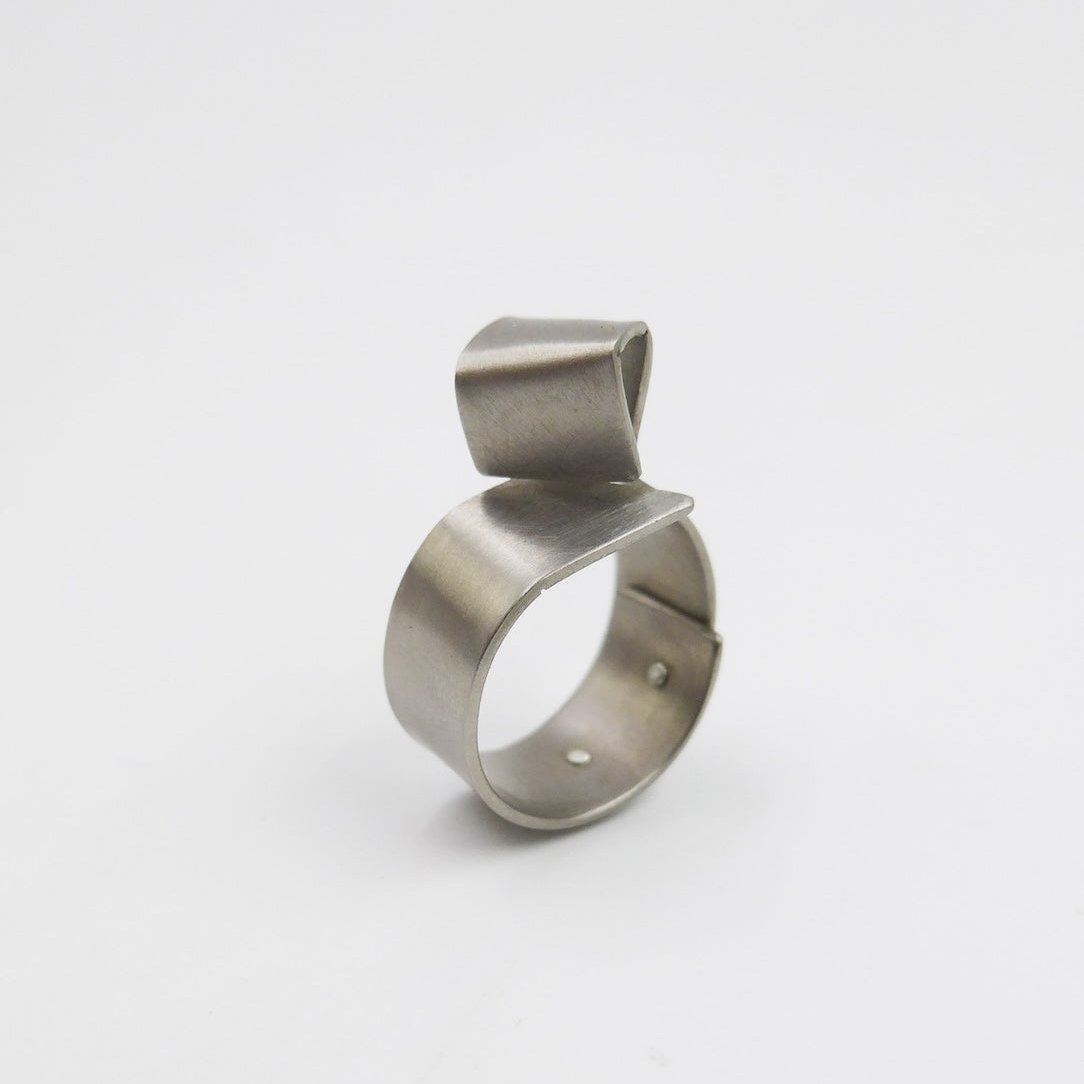 Bague sculpturale construite avec deux plaques de titane grises rivetées l'une à l'autre. Le métal s'enroule autour du doigt comme un anneau large. Sur le dessus le  titane est plié en forme de cube irrégulier.