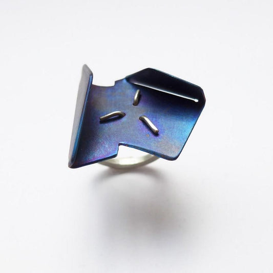 Bague géométrique réalisée avec une plaque de titane bleuÏ à la flamme, dont les deux côtés opposés sont légèrement replier. Ce bijou est fixé au centre par trois griffes sur un anneau en argent.