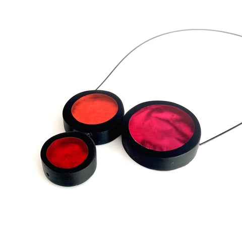 Détail, collier 3 ronds de résine couleurs rouge, orange et fushia assemblés sur un câble noir aimanté.