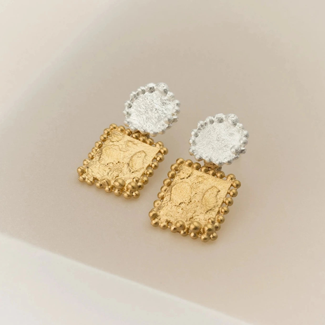 Ces boucles d'oreilles se composent de deux éléments distincts. Sur l'oreille, un disque en argent. En suspension, un carré en argent plaqué or jaune. La granulation délicate souligne les détails du métal travaillé. Décor beige.