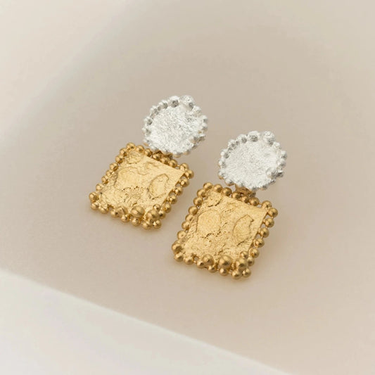 Ces boucles d'oreilles se composent de deux éléments distincts. Sur l'oreille, un disque en argent. En suspension, un carré en argent plaqué or jaune. La granulation délicate souligne les détails du métal travaillé. Décor beige.