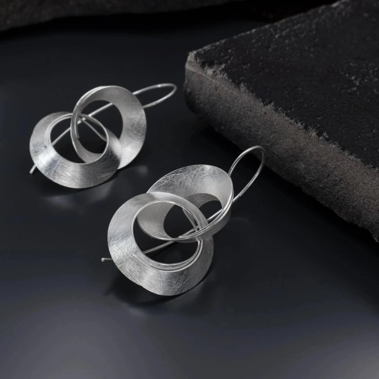 Grandes boucles d'oreilles en argent composés de deux anneaux entrelacés, montés sur deux longues tiges forme crochet pour fermeture. bijoux sur fond noir.