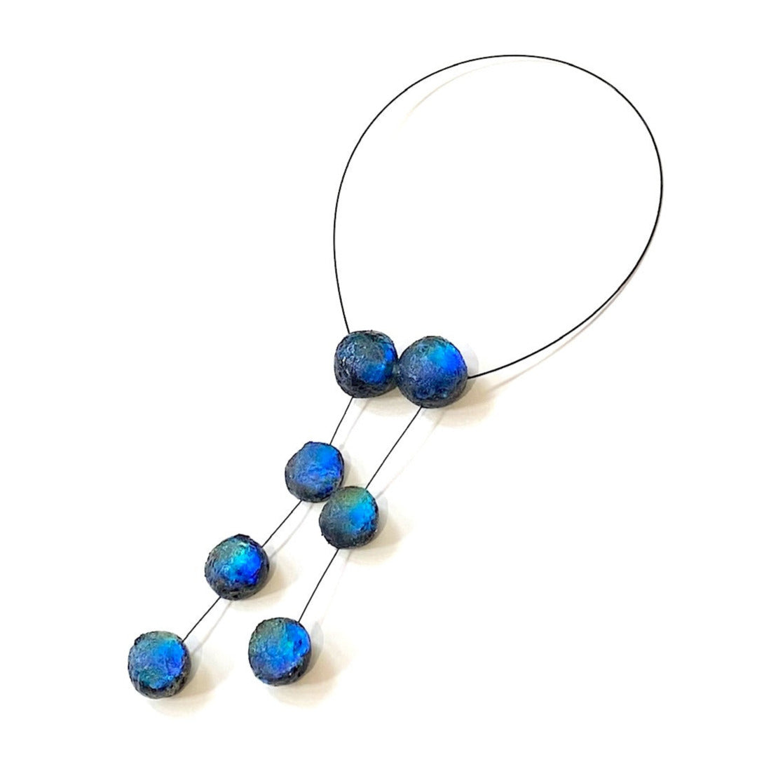 Collier en forme de cravate, composé de 7 sphères en résine aux nuances de bleu, aux reflets irisés, assemblées sur un cable noir. Ce bijou asymétrique capte la lumière à chacun de vos mouvements. 