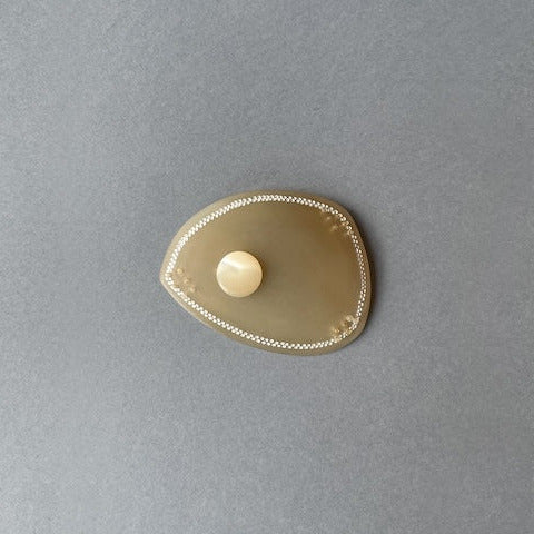 Broche en corne dorée, ornée d’une délicate frise composée de deux rangées de petits points blancs peints à la main. Une pastille de nacre ivoire apporte lumière et éclat.