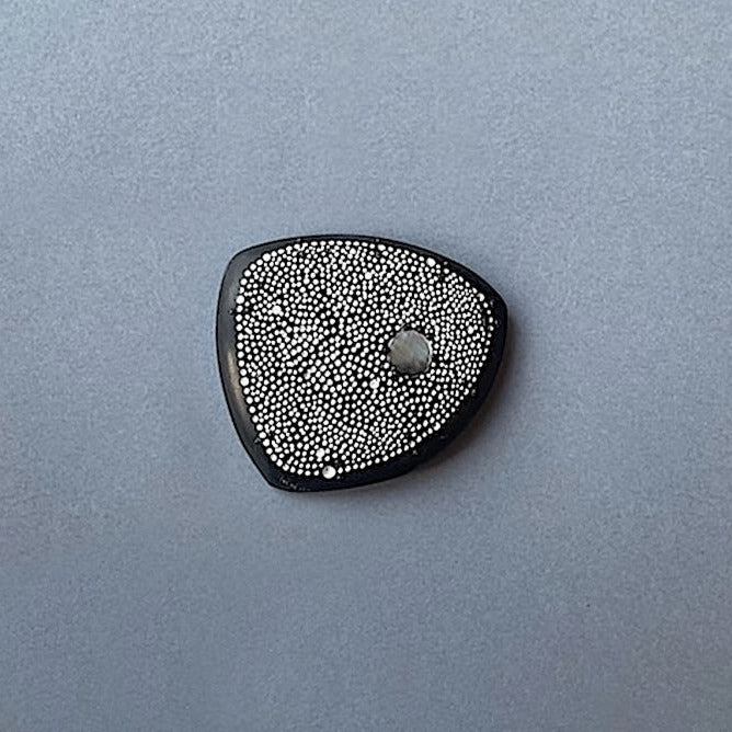 La broche Étoilée en corne noire sculptée et patinée, ornée d’une pastille de nacre grise. En forme de galet, elle est décorée  de délicats points blancs peints à la main, elle évoque la finesse du pointillisme et rend hommage à l'art aborigène.