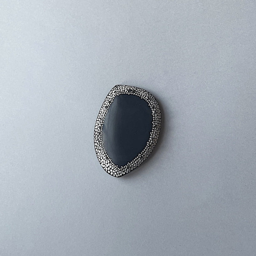 Broche unique en corne noire ornée d'une frise de 50 mm composée de points blancs peints à la main, rappelant le pointillisme de l'art aborigène. De mini perles d'hématites soulignent ce travail abstrait. 