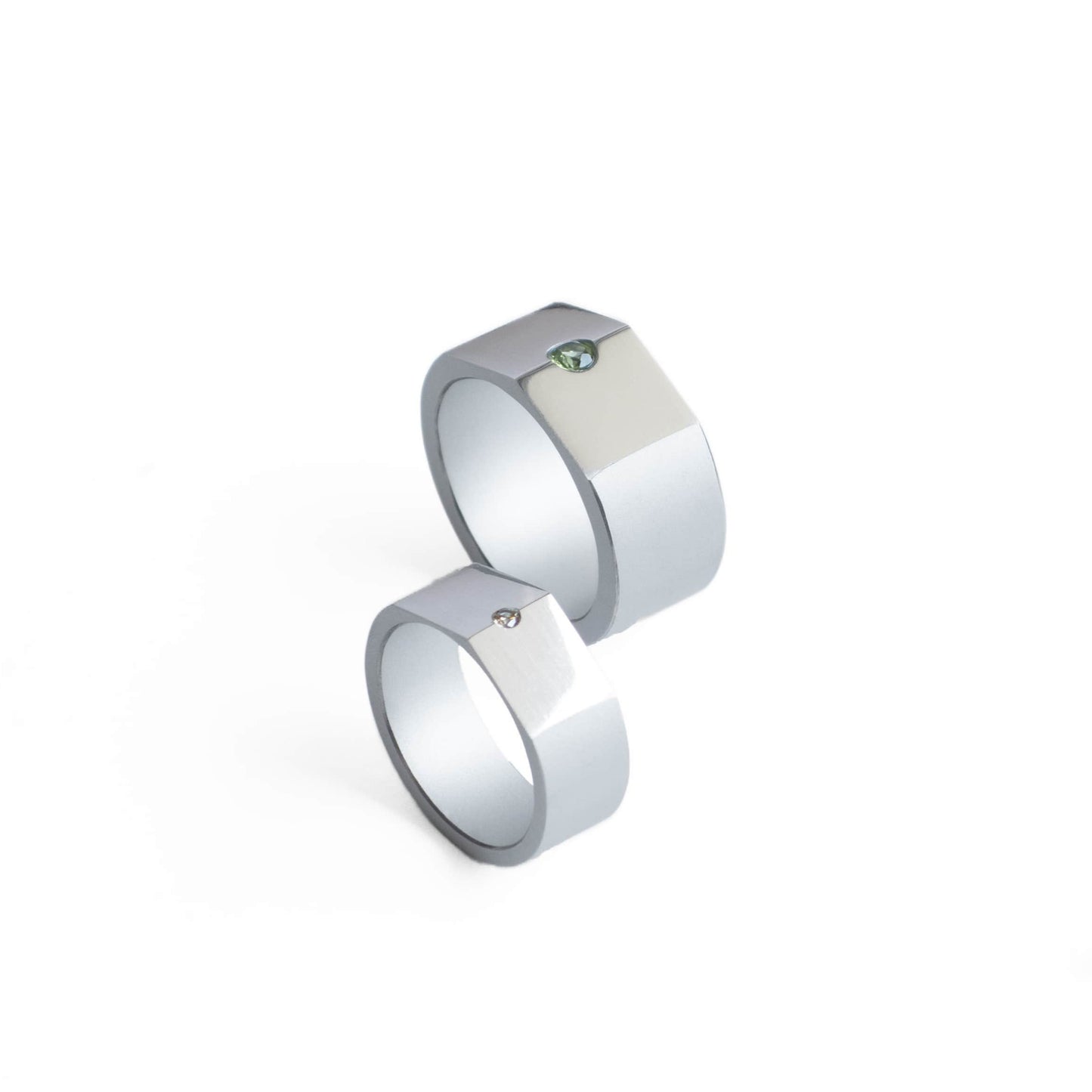 Deux bague de différentes tailles sont présentées debout l'une devant l'autre. En aluminium anodisé gris, l'une est ornée d'un péridot, l'autre d'un diamant.