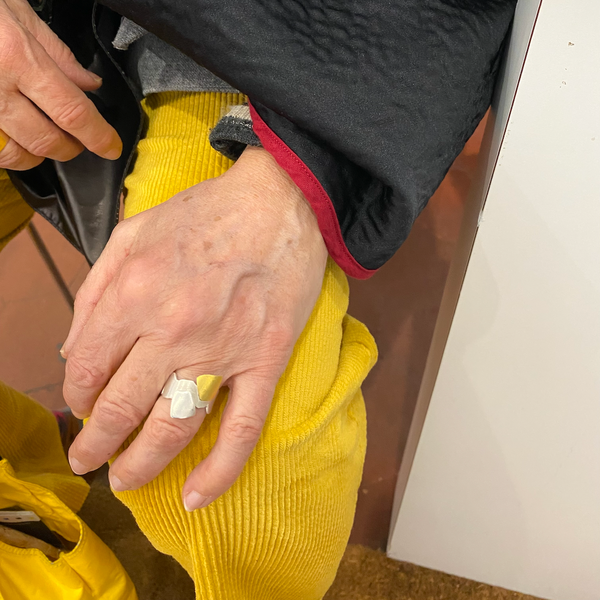 Bague géométrique en argent brossé formée de carrés irréguliers et d'un carré en or jaune; Bijou porté, main posée sur genoux, pantalon jaune.