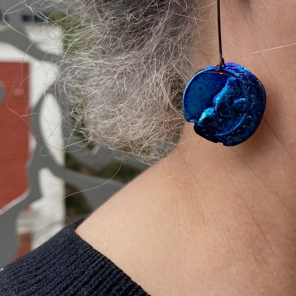 Boucle pendante en titane bleu et texturé en forme de disque irrégulier présenté sur une tige droite en titane noir. Détail profil et cou.