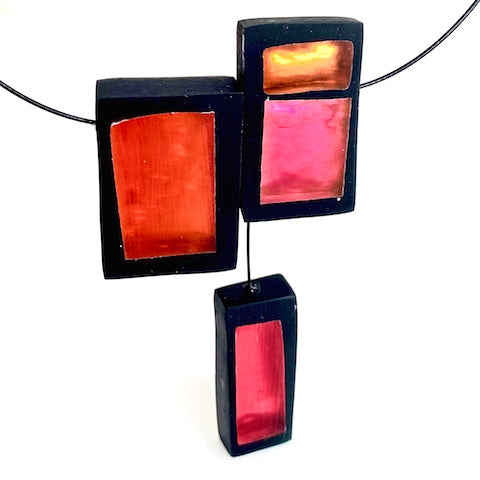 Collier avec trois rectangles de résine rouge, orange et rose assemblés sur un câble noir. Fermoir aimant inséré dans la résine.