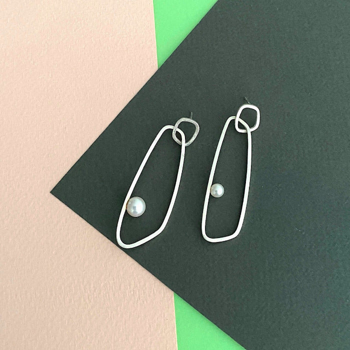 Boucles d'oreilles en argent de type mobile composées de deux pièces ornées d'une perle de culture blanche, posées sur fond de couleurs rose, vert, gris