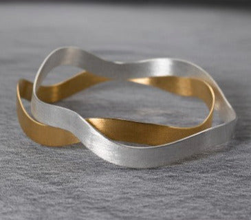 Deux bracelet ondulés, un bracelet en argent brossé, l'autre en argent plaqué or jaune, posés sur fond gris
